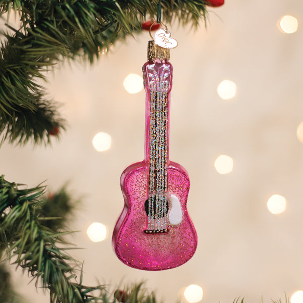Pink glittery guitar ornament hanging on a Christmas tree with lights in the background.