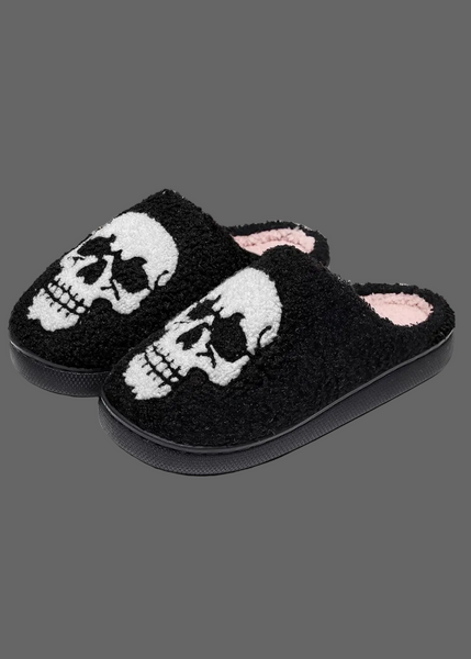 A pair of black fuzzy slip-on slippers featuring a large white skull design on the top, with a light pink lining visible inside.
