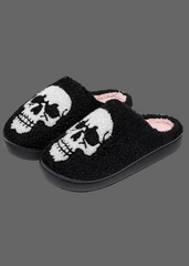 A pair of black fuzzy slip-on slippers featuring a large white skull design on the top, with a light pink lining visible inside.