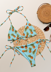 Tropical-themed bikini set with pineapple pattern, straw bag, and sunglasses on a light background