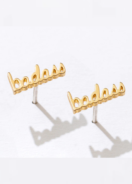 Gold 'badass' earrings on a stand with a white background
