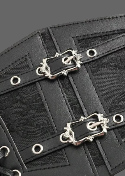 Close-up of a black leather accessory with two ornate silver-tone decorative buckles, silver grommets, and a textured patterned fabric background.