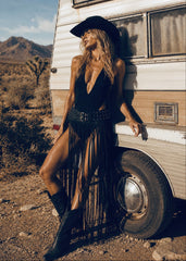Woman in a black fringe dress and hat standing next to a vintage camper in a desert setting