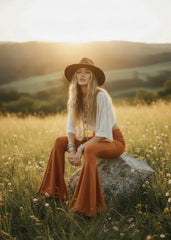 Model seated on a rock wearing rust bell bottom pants with wide flared hems and a cream cropped top.