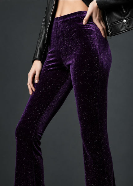 A close-up of a model wearing high-waisted, flared purple velvet pants with a subtle glitter texture, styled with a black leather jacket against a dark gray studio background.