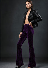 A model wearing high-waisted, flared purple velvet pants with a subtle sequin texture, styled with a black leather moto jacket and black heels against a dark gray studio background.