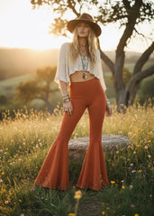 Model wearing high-waisted rust bell bottom pants with a cream cropped top and brown hat in a sunlit meadow